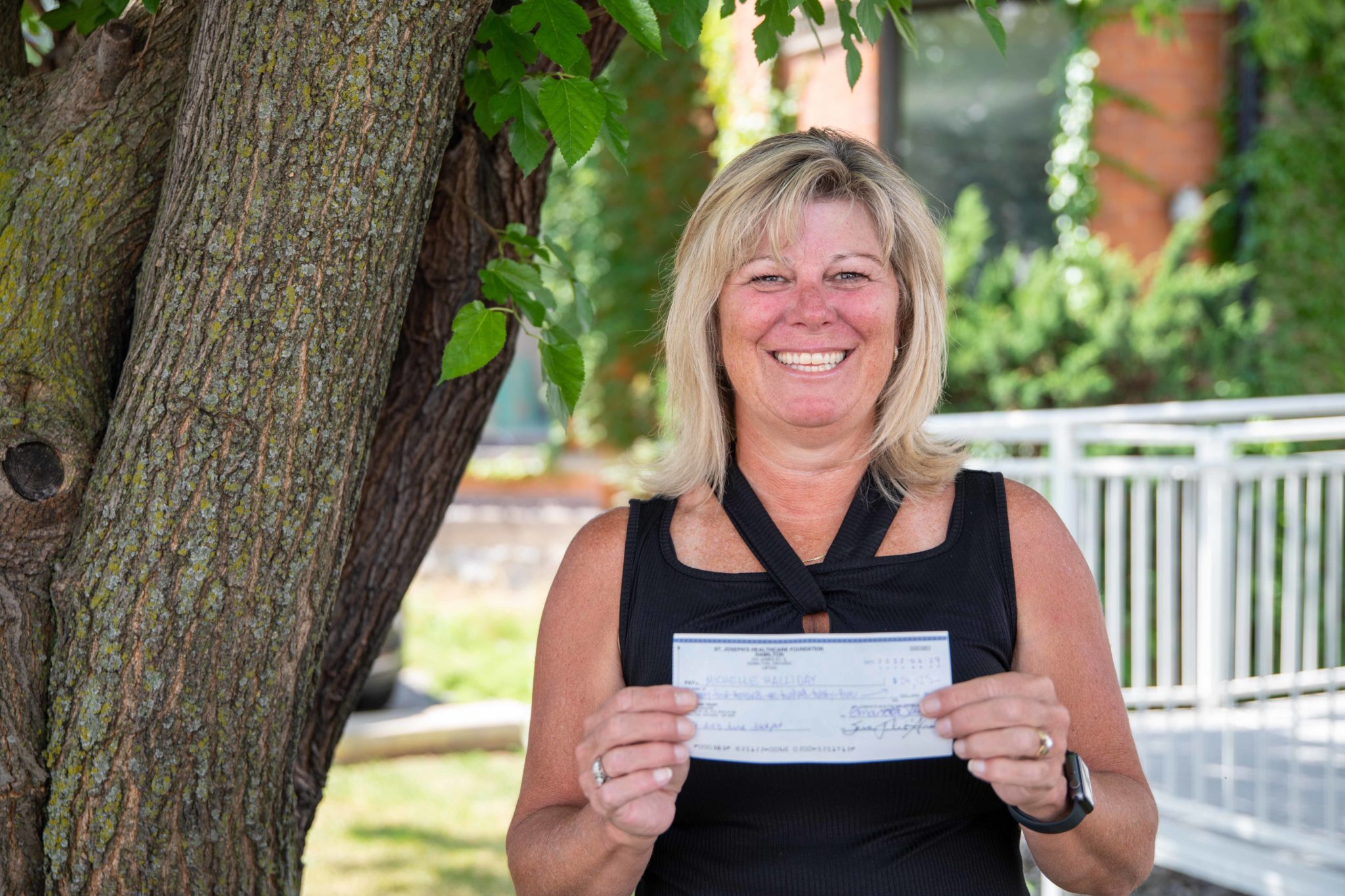 St. Joe's June Jackpot lottery winner, Michelle H., is photographed outdoors next to a large tree. She smiles while holding up her winning cheque.